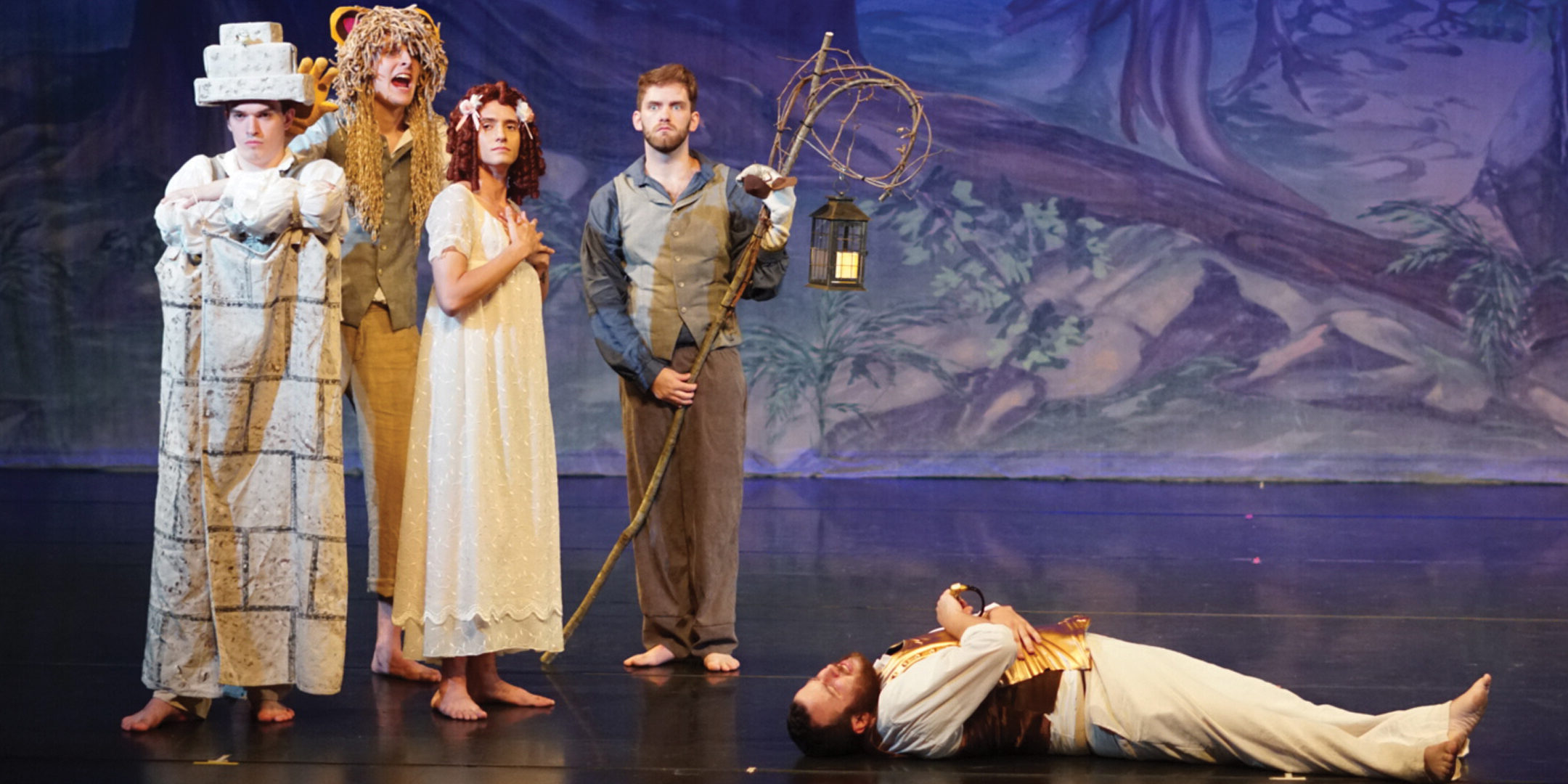 Four actors stand over a reclining figure on stage.