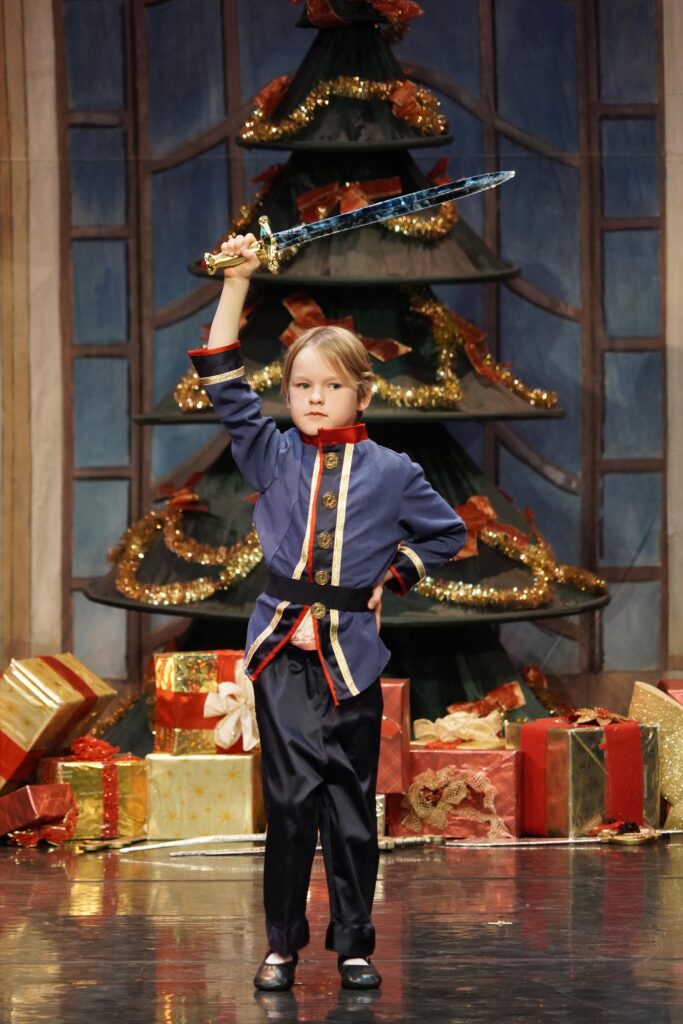A young dancer in the Nutcracker holds up a sword in front of the Christmas tree.