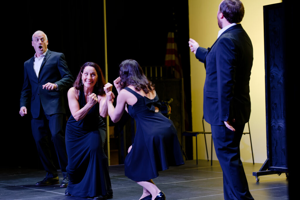 Two women playfully hold up fists on stage, while two men react.