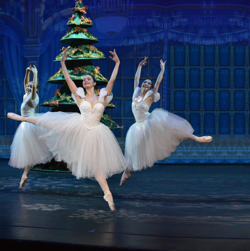 Three dancers in white costumes dance in front of a Christmas tree