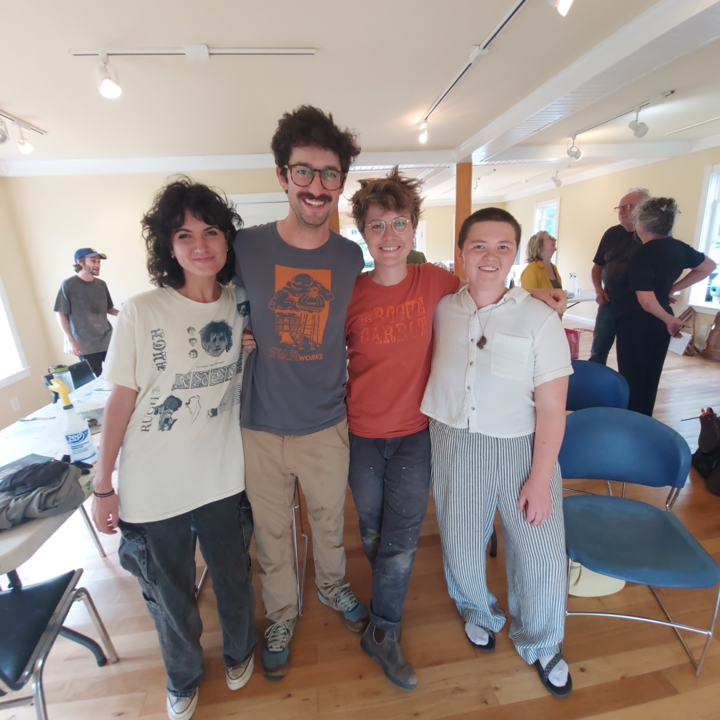 A group of four people stand together with their arms around each other in an art studio