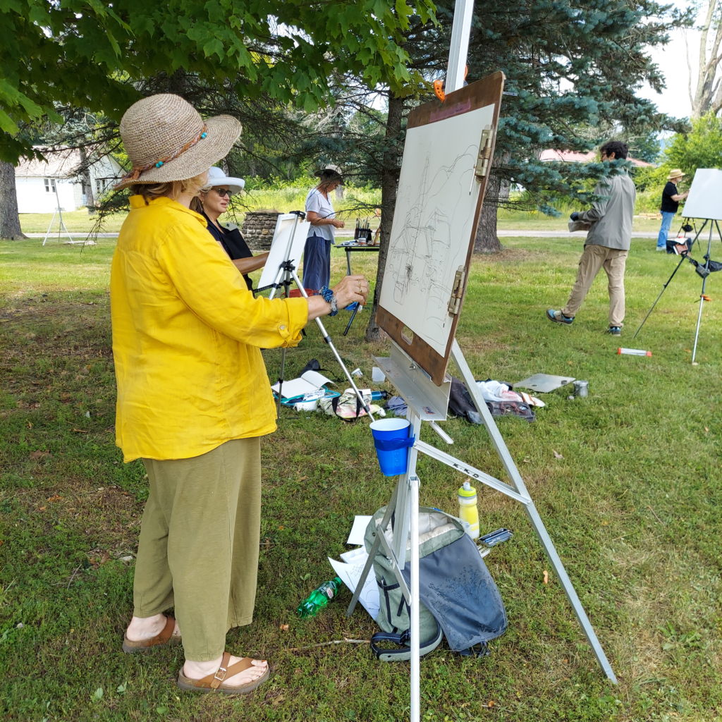 A woman in a hat and yellow shirt stands in the foreground in front of an easel, painting outside.