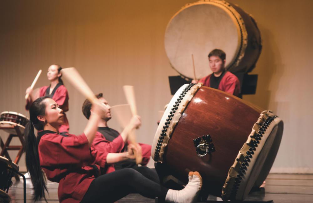 A group of people sit on a stage playing large taiko drums.