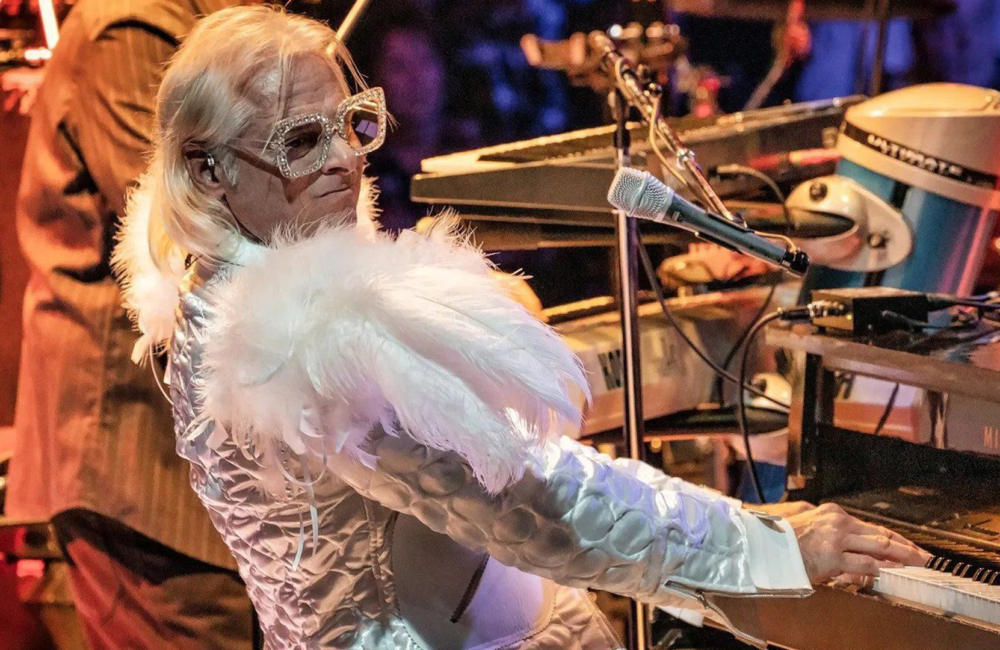 Man dressed in a flamboyant white costume with sparkling sunglasses plays a piano.