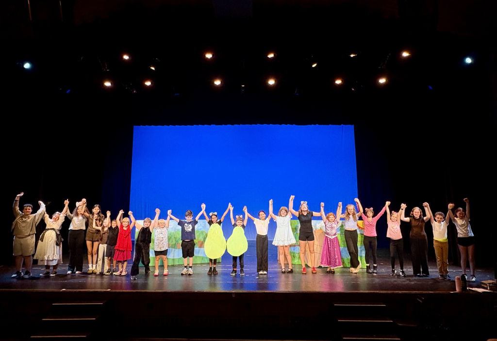 A group of kids in colorful costumes raise their hands on stage.